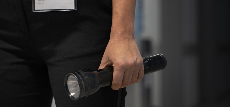 Mano di un addetto in divisa nera con badge al petto che impugna una torcia metallica scura in un corridoio interno