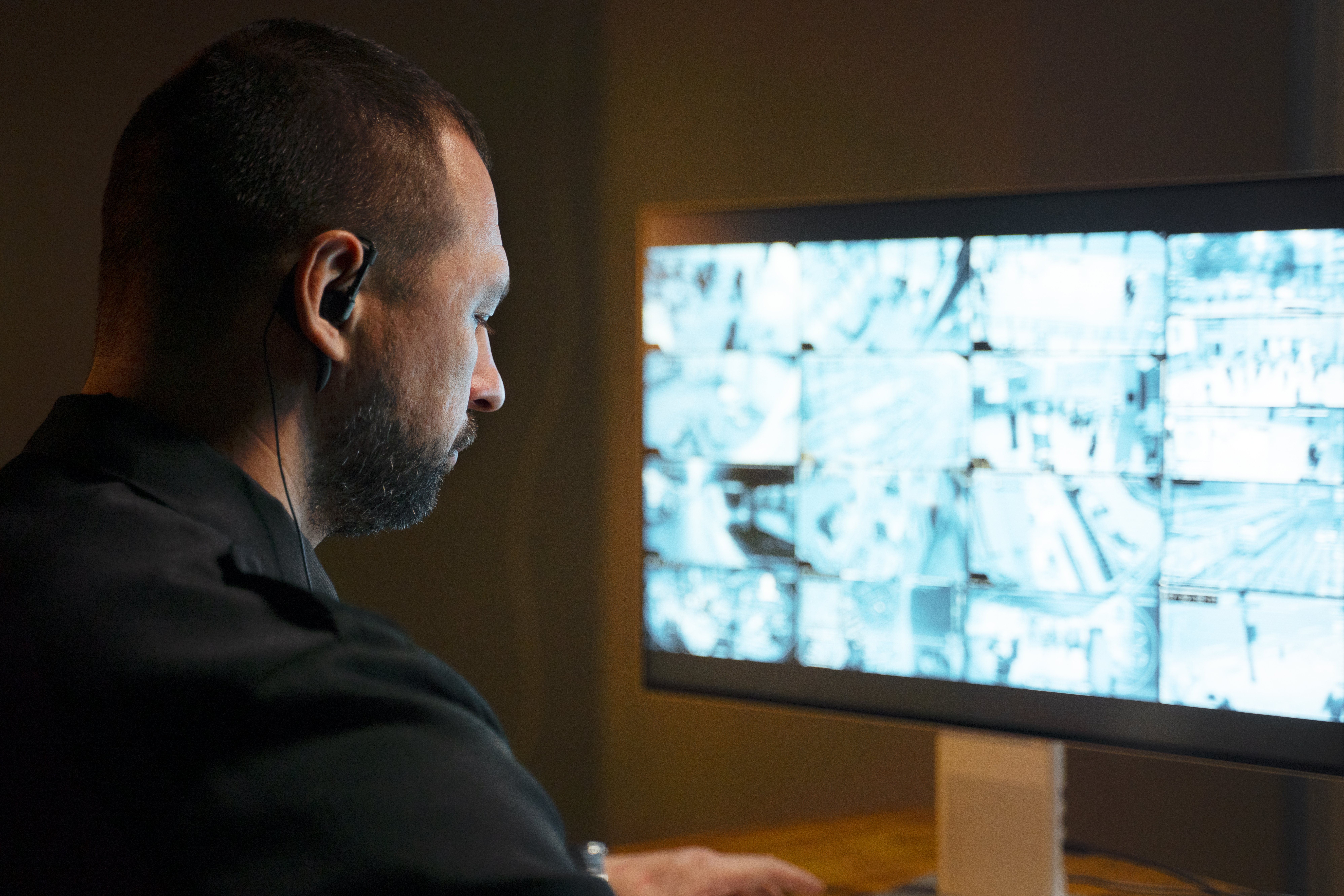 Operatore con auricolare e divisa scura osserva un monitor con griglia di immagini delle telecamere in una stanza in penombra